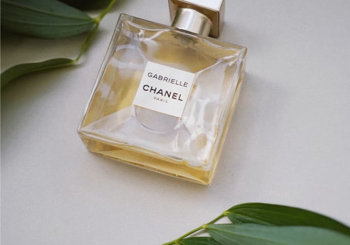 A bottle of Gabrielle Essence perfume laying flat on a white table with green leaves above and below it.